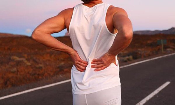 An athlete on an open road at dusk, hands resting on a sore lower back beneath a peach-and-blue sky.